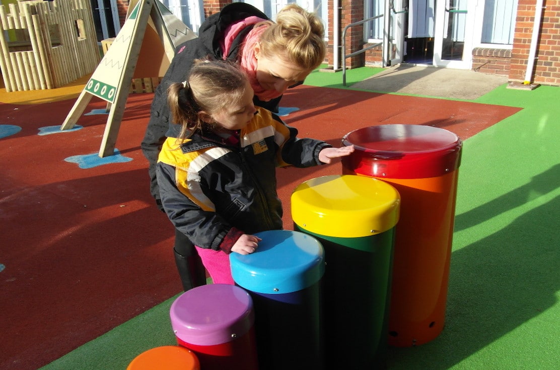 a child using their hand to play multicoloured bongo drums being assisted by an adult