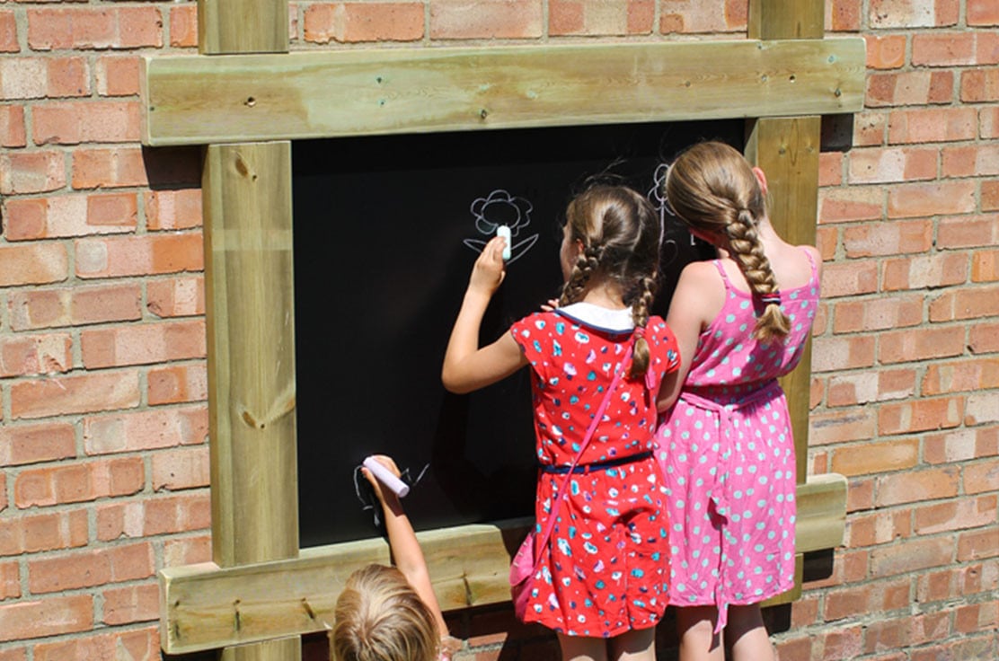 three children using white chalk to draw flowers on a chalkboard fixed to a brick wall