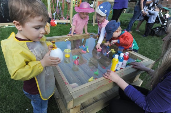 four children using primary colour paint and magnetic letters to create images on a wooden frame with a perspex table top