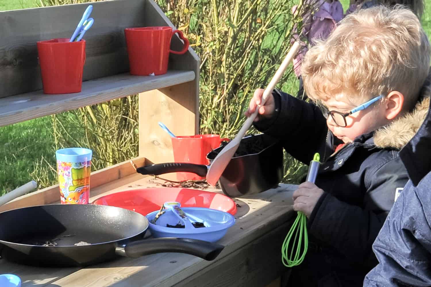 a child holding a green whisk and wooden spoon pretending to cook with a frying pan and saucepan on a wooden mud pie kitchen
